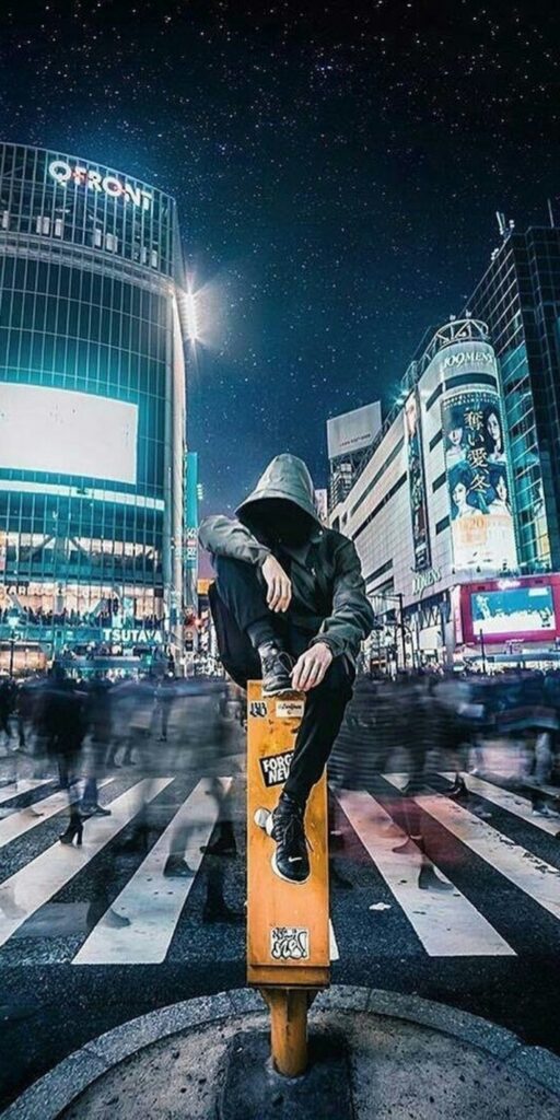 A person in a hoodie sits on a pole at a busy, illuminated city intersection at night. Blurred pedestrians cross the street, and bright billboards light up the background with a starry sky above.
