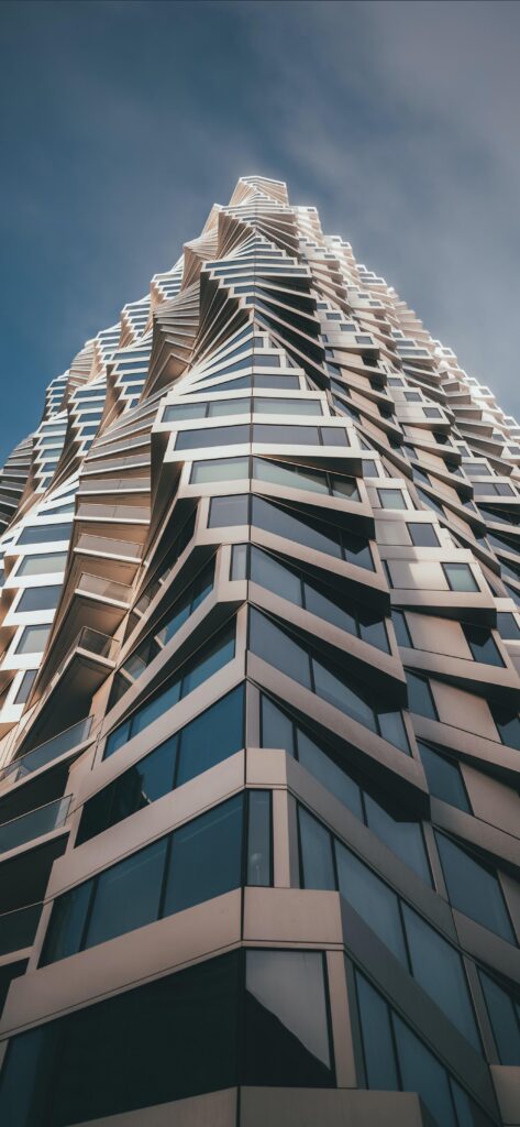 A modern skyscraper with a unique, twisting architectural design, featuring numerous glass windows and angled surfaces, against a clear sky. The perspective is from the base of the building looking upward.
