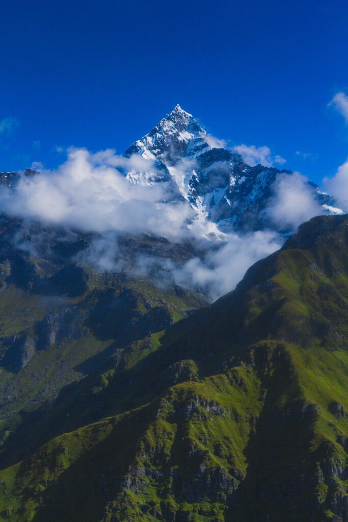 A majestic snow-capped mountain peak rises above lush green hills under a clear blue sky. White clouds partially veil the mountain, creating a dramatic and picturesque landscape.
