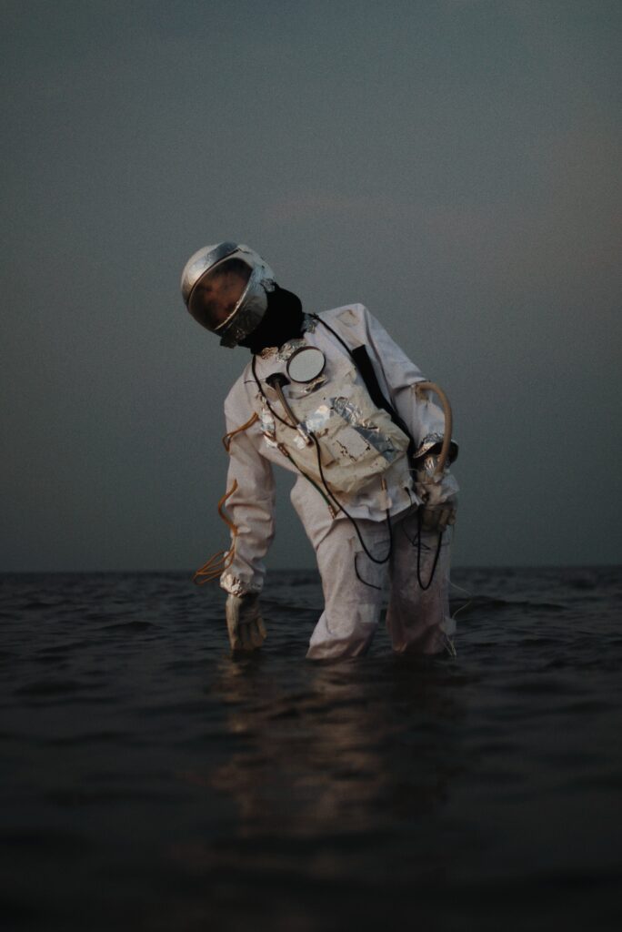 A person wearing a space suit stands partially submerged in water against a dark, cloudy sky. The suit appears vintage with reflective surfaces and a helmet. The scene has a mysterious and surreal atmosphere.
