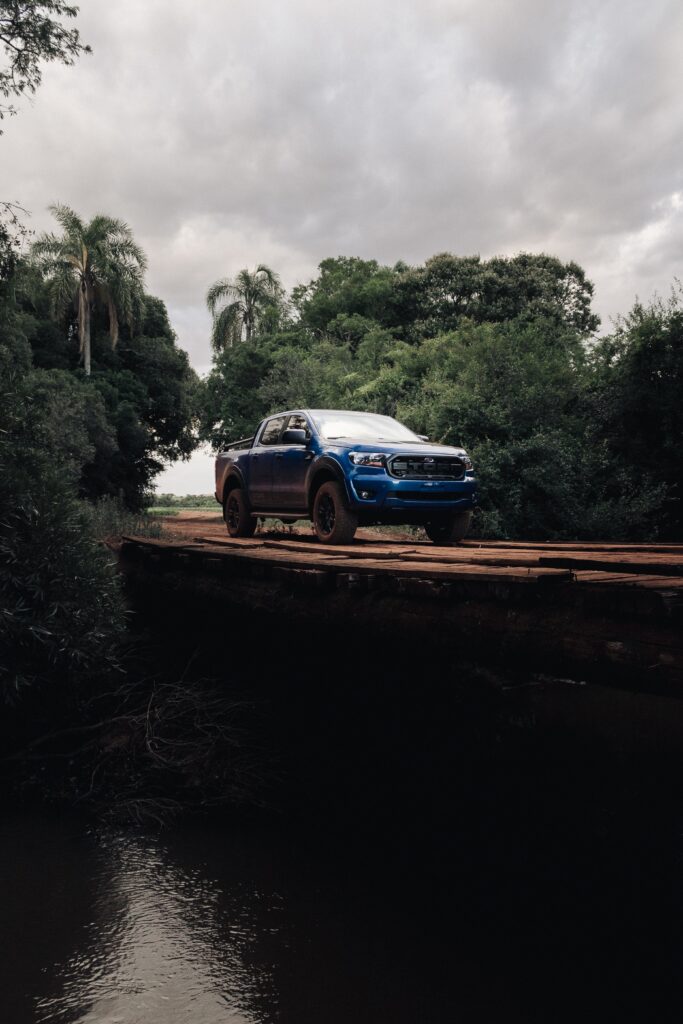 A blue pickup truck on a narrow, rusty bridge surrounded by dense green foliage and trees, with a cloudy sky overhead. The scene conveys a sense of adventure in a remote, natural setting.
