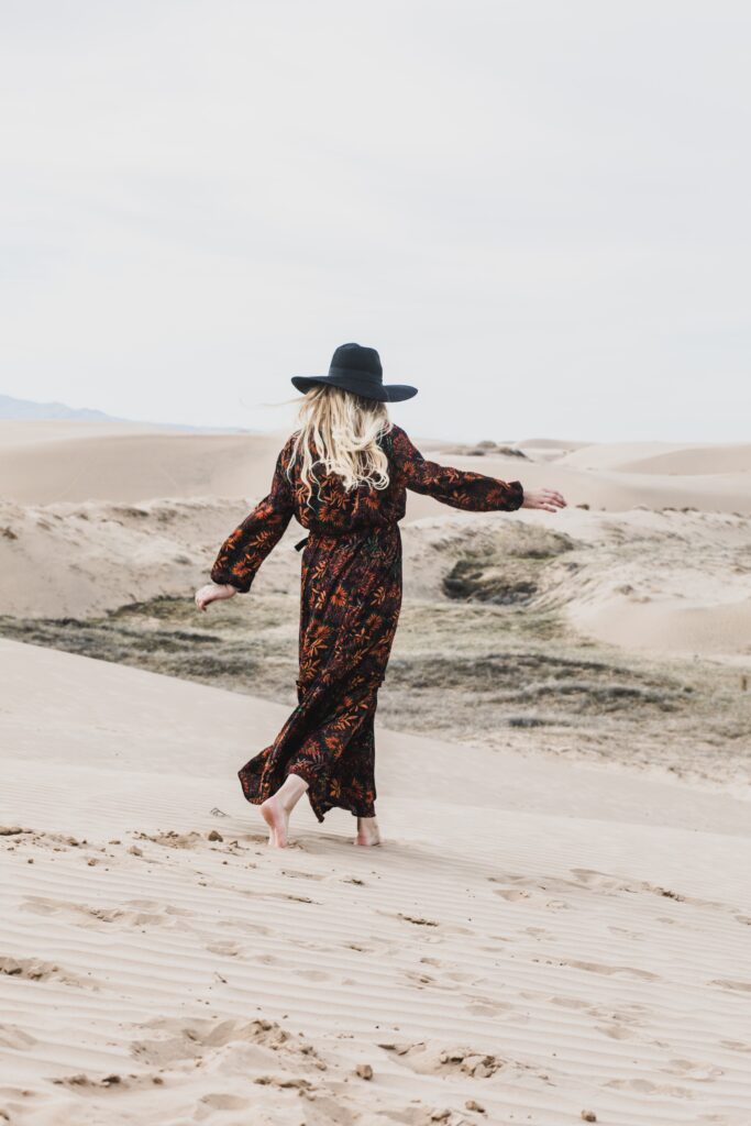 A woman in a long, patterned dress and black hat walks barefoot on a sandy desert with gentle dunes. She walks away from the camera, her arms slightly outstretched. The sky is overcast, adding a serene atmosphere to the scene.
