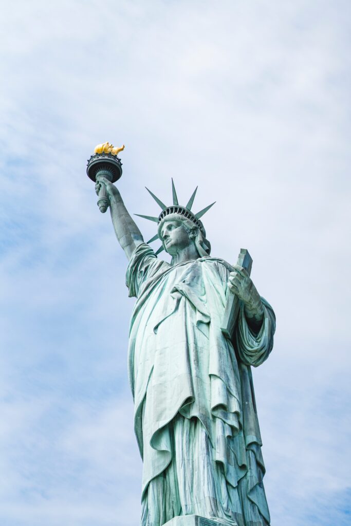 The Statue of Liberty stands tall against a blue sky, holding a torch in her raised right hand and a tablet in her left. Her distinctive crown with seven spikes is visible.
