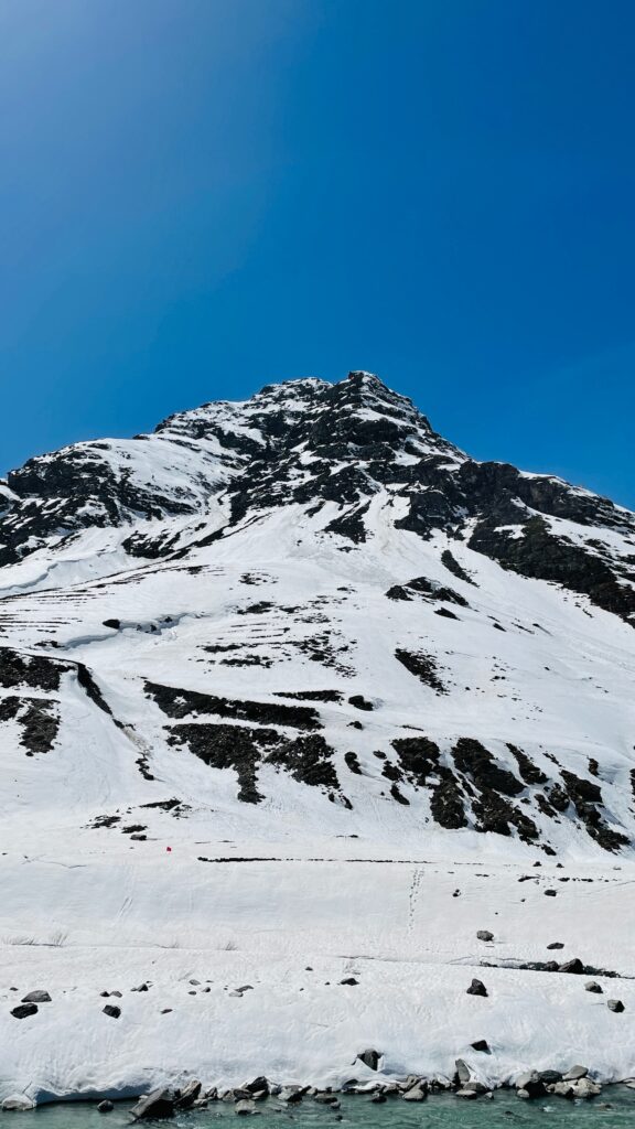 Snow-covered mountain peak against a clear blue sky, with rocky patches visible. Snow blanketed foreground with scattered rocks, creating a serene winter landscape.
