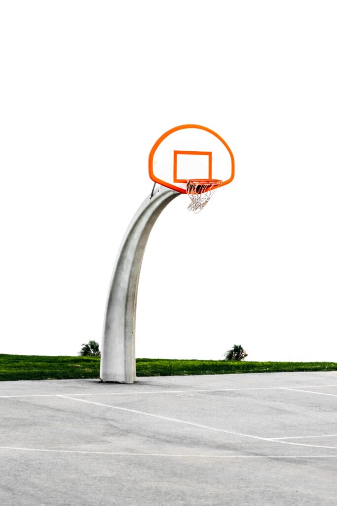 A solitary outdoor basketball hoop with an orange rim and a white backboard stands on a concrete court. The sky is a clear white, and there are small patches of green grass visible in the background.
