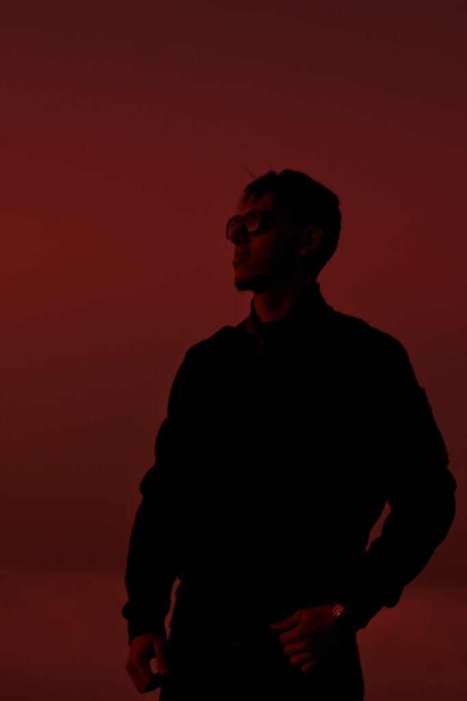 A person wearing sunglasses stands in silhouette against a vibrant red-orange background, creating a dramatic and mysterious atmosphere.
