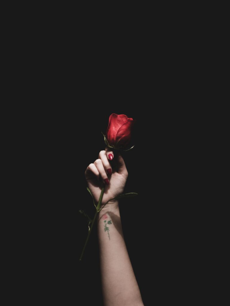 A hand with red painted nails holds a single red rose against a dark background. A small rose tattoo is visible on the wrist. The composition is minimalistic, emphasizing the contrast between the vibrant rose and the darkness.
