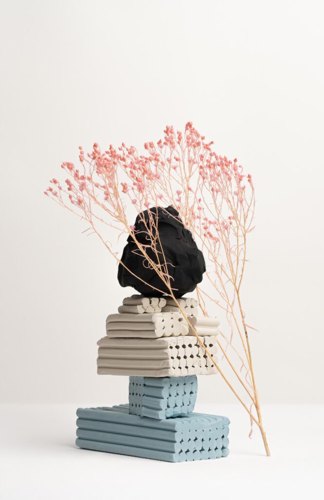 A minimalist sculpture with a stack of rectangular, textured blocks in blue, grey, and beige, topped by a black sphere. Delicate pink dried flowers are artfully arranged around it, set against a plain white background.
