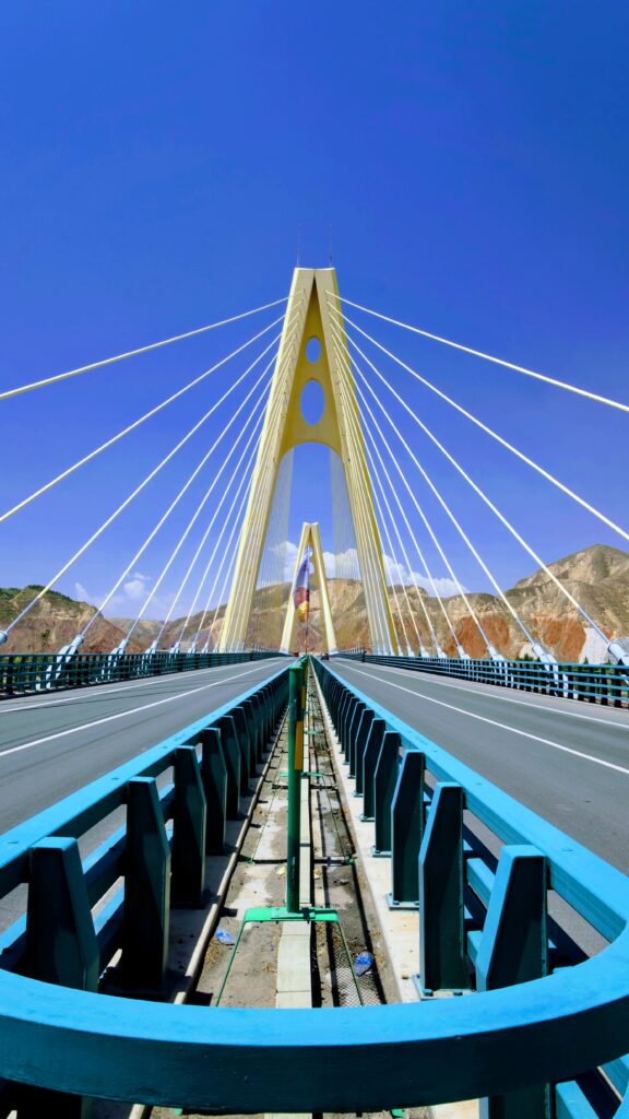 A dramatic view of a modern suspension bridge with a central yellow tower and white cables against a clear blue sky. The road leads directly toward the tower, and mountains are visible in the background.
