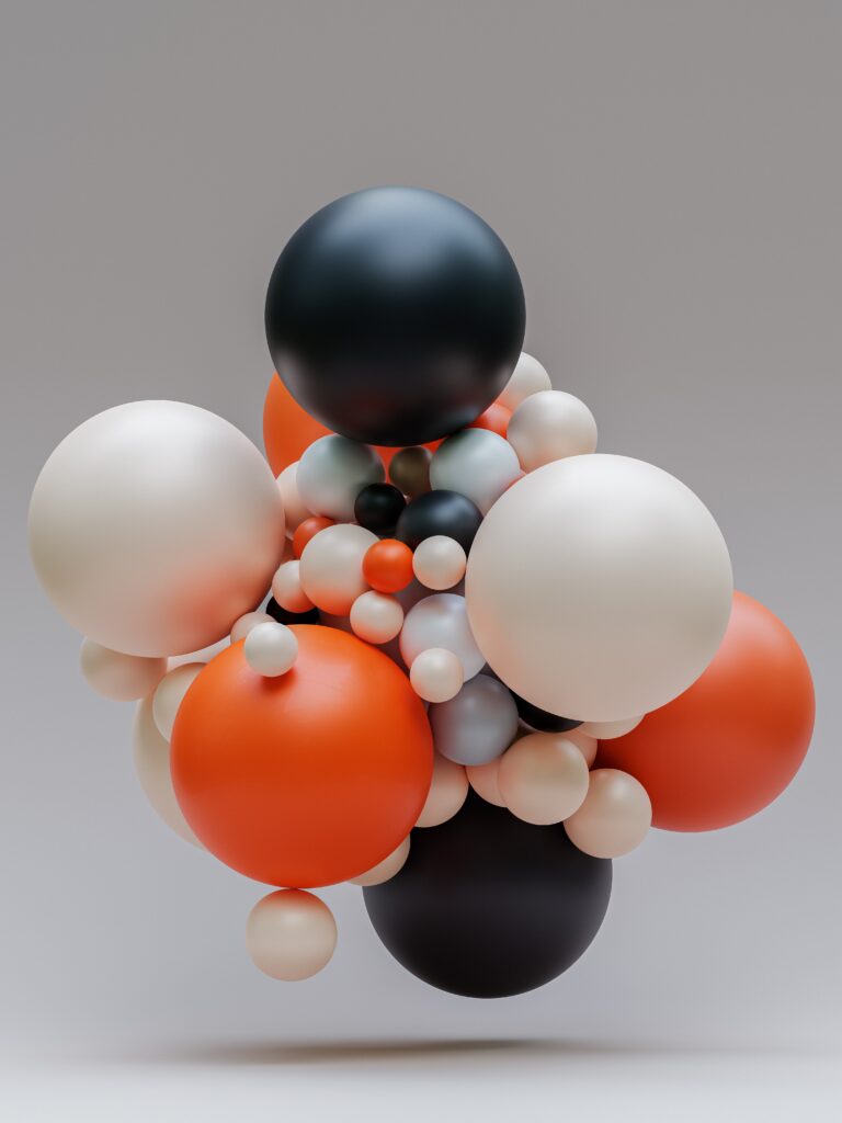 A cluster of multicolored balloons, including orange, white, black, and gray, of various sizes floating against a plain gray background. The arrangement creates a visually striking and balanced composition.
