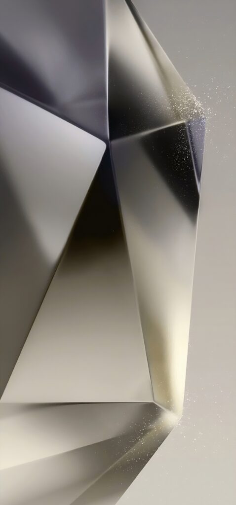 Close-up of a faceted, metallic geometric object with smooth surfaces reflecting light. The background is a gradient of gray tones, creating a modern and sleek appearance. Small specks of light appear near the edges.
