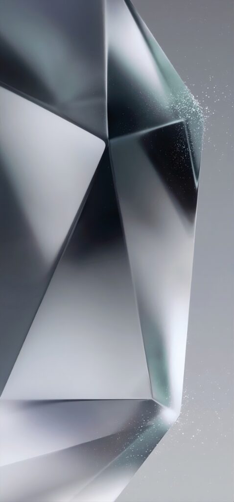 Close-up of a faceted, geometric object with a metallic sheen, set against a gradient gray background. Small particles or dust appear to be scattered around the right side, enhancing the reflective and textured surface.
